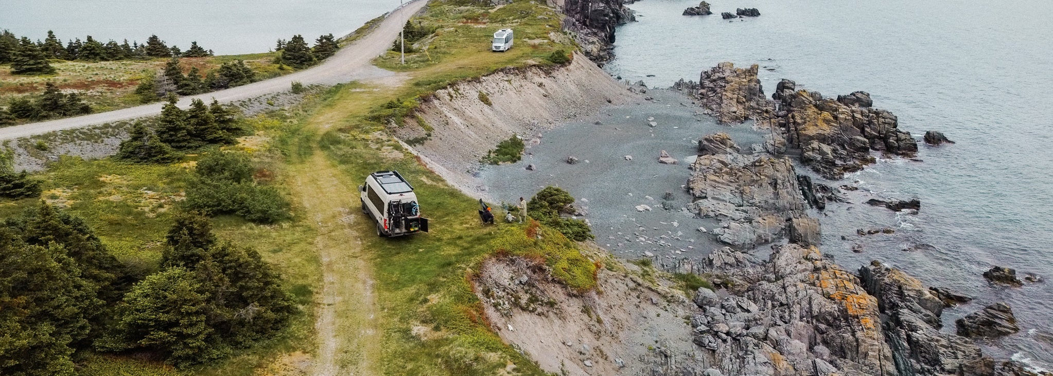 Van parked on a grassy area near a rocky coastline with water and mountains in the background.